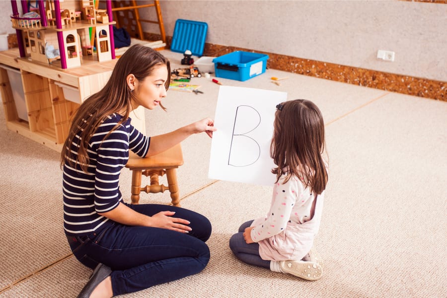 Profesional de la logopedia muestra una tarjeta con la letra B a una niña sentada en alfombra de juego, sesión de estimulación articulatoria y del lenguaje en casa.